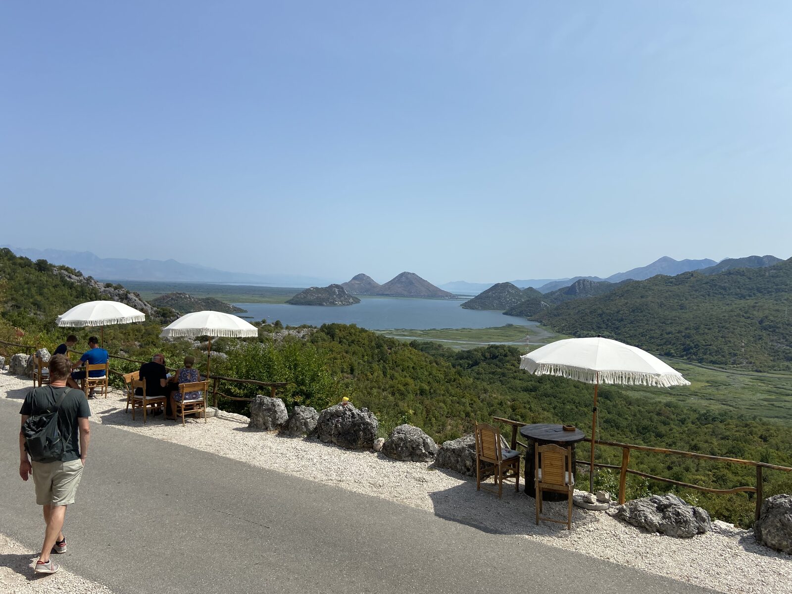 Visitors at a shaded lookout terrace above Lake Skadar, with forested hills and conical islands rising from the calm water